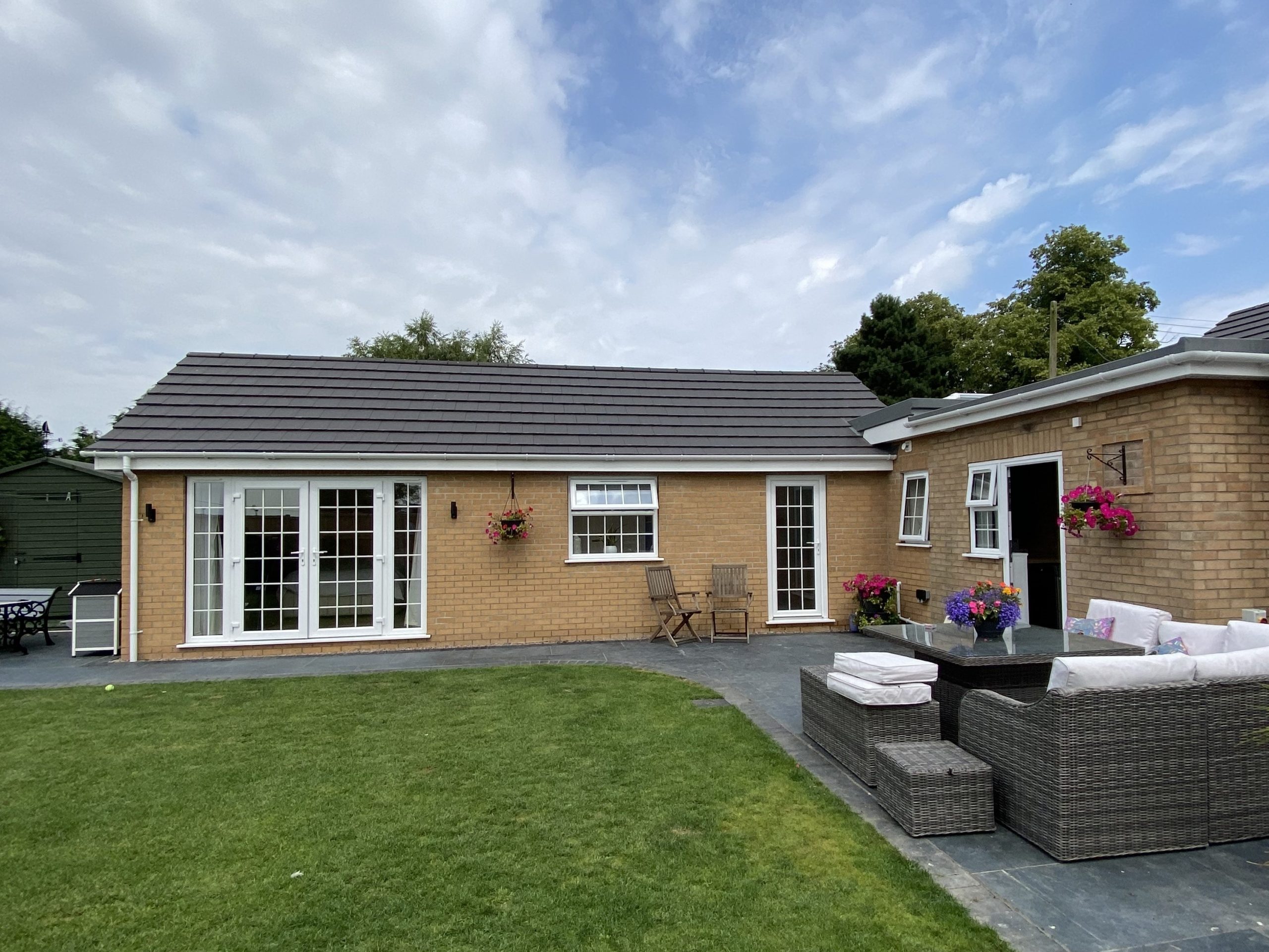 Single-story brick house with gray roof, white-framed doors and windows, outdoor seating area with wicker furniture, hanging flower baskets, and a well-kept lawn under a partly cloudy sky.