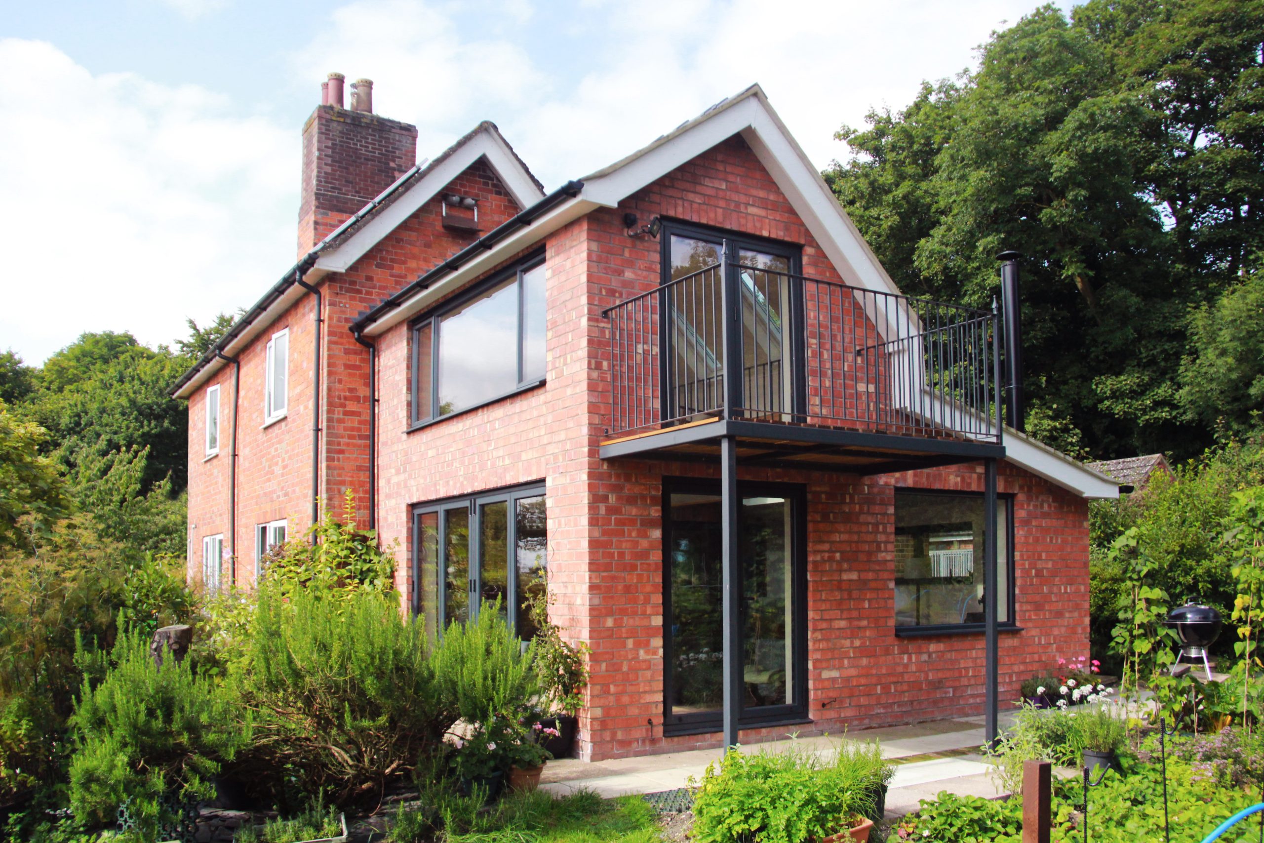A two-story red brick house with large windows, a small balcony, and a garden in the front, surrounded by greenery and trees under a partly cloudy sky.