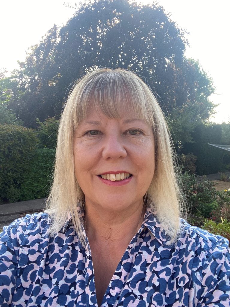 A smiling woman with straight, shoulder-length blonde hair stands outdoors in a garden, wearing a blue and white patterned shirt. Elegant architecture peeks through the lush bushes and trees visible in the background.
