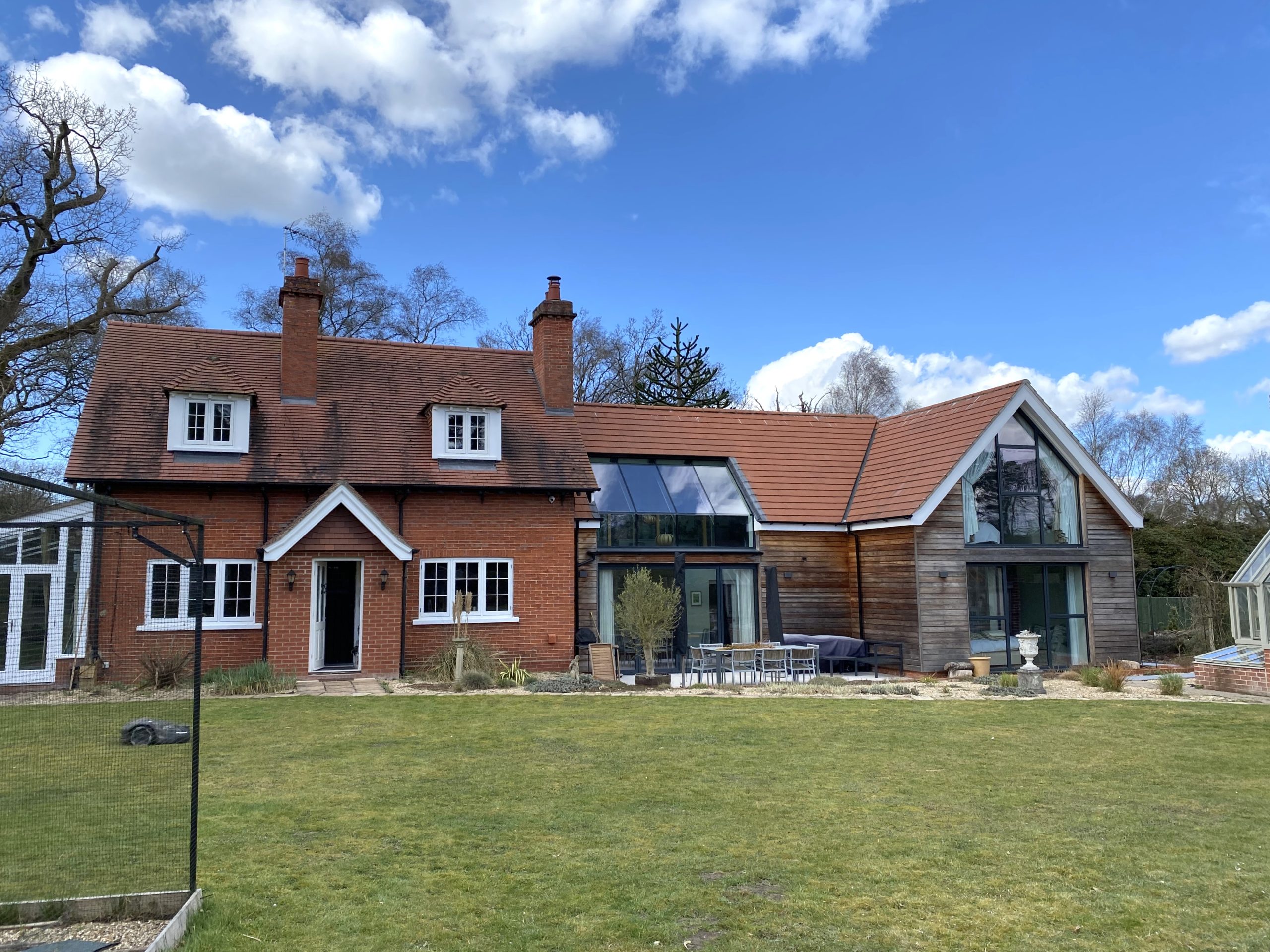 A red-brick house with a pitched roof and modern glass extension, set in a grassy yard under a blue sky with clouds.