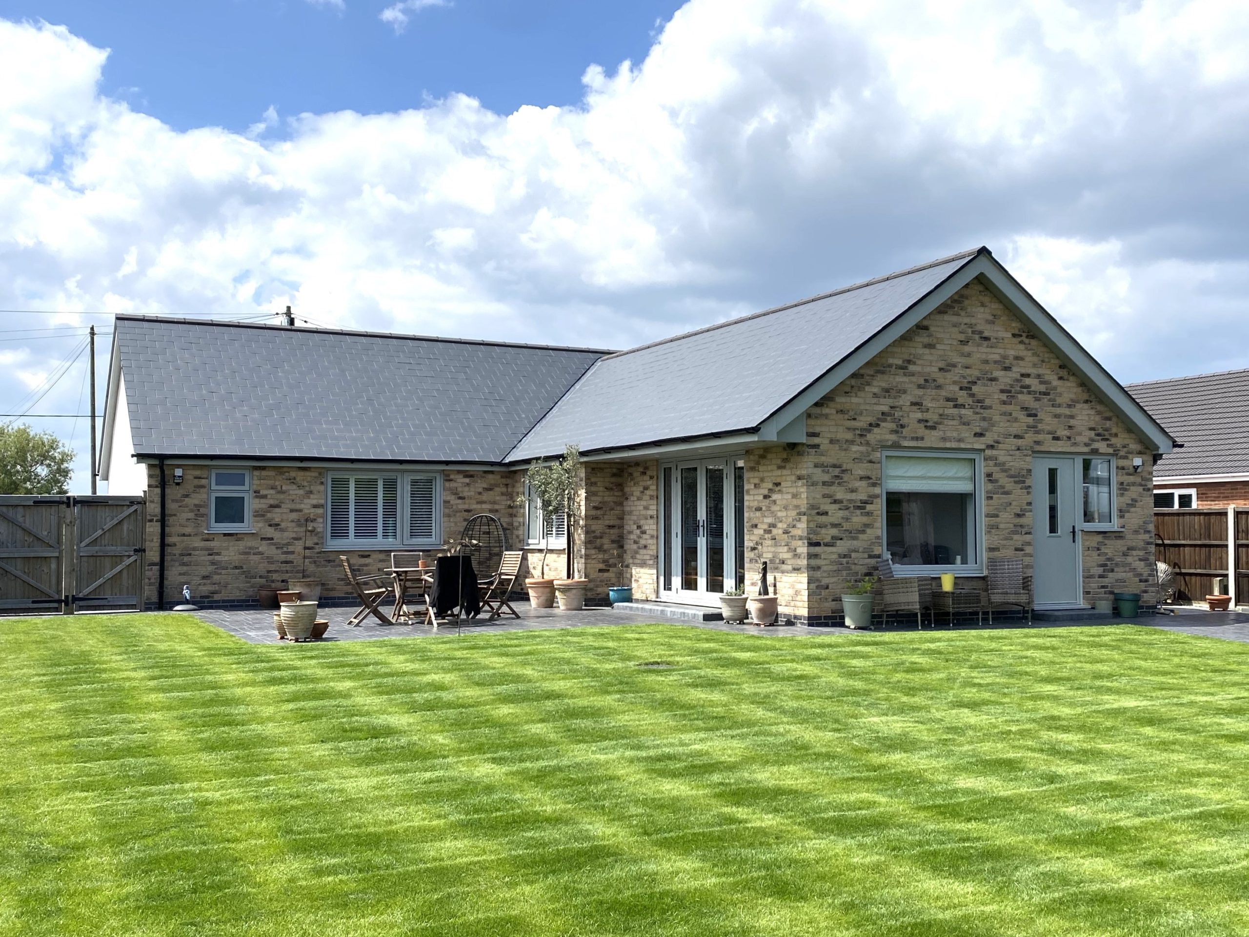 A modern single-story brick house with a gray roof, large windows, and a patio area with outdoor seating. The house is surrounded by a neatly mowed, green lawn under a partly cloudy sky.