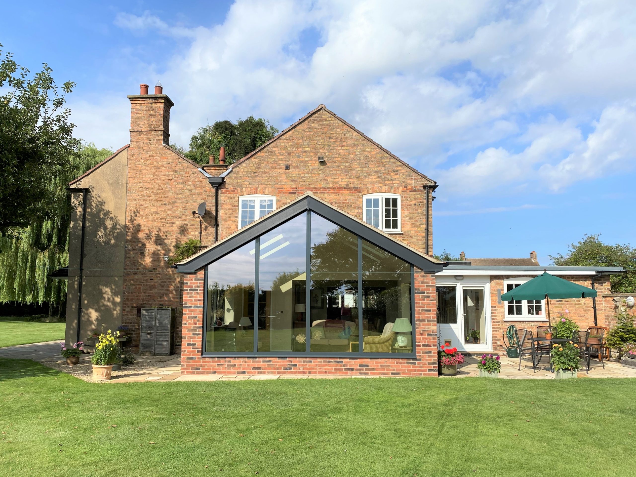 A brick house with a modern glass extension, surrounded by a green lawn. Outdoor seating with a green umbrella is on a patio to the right. Trees and blue sky with clouds are in the background.