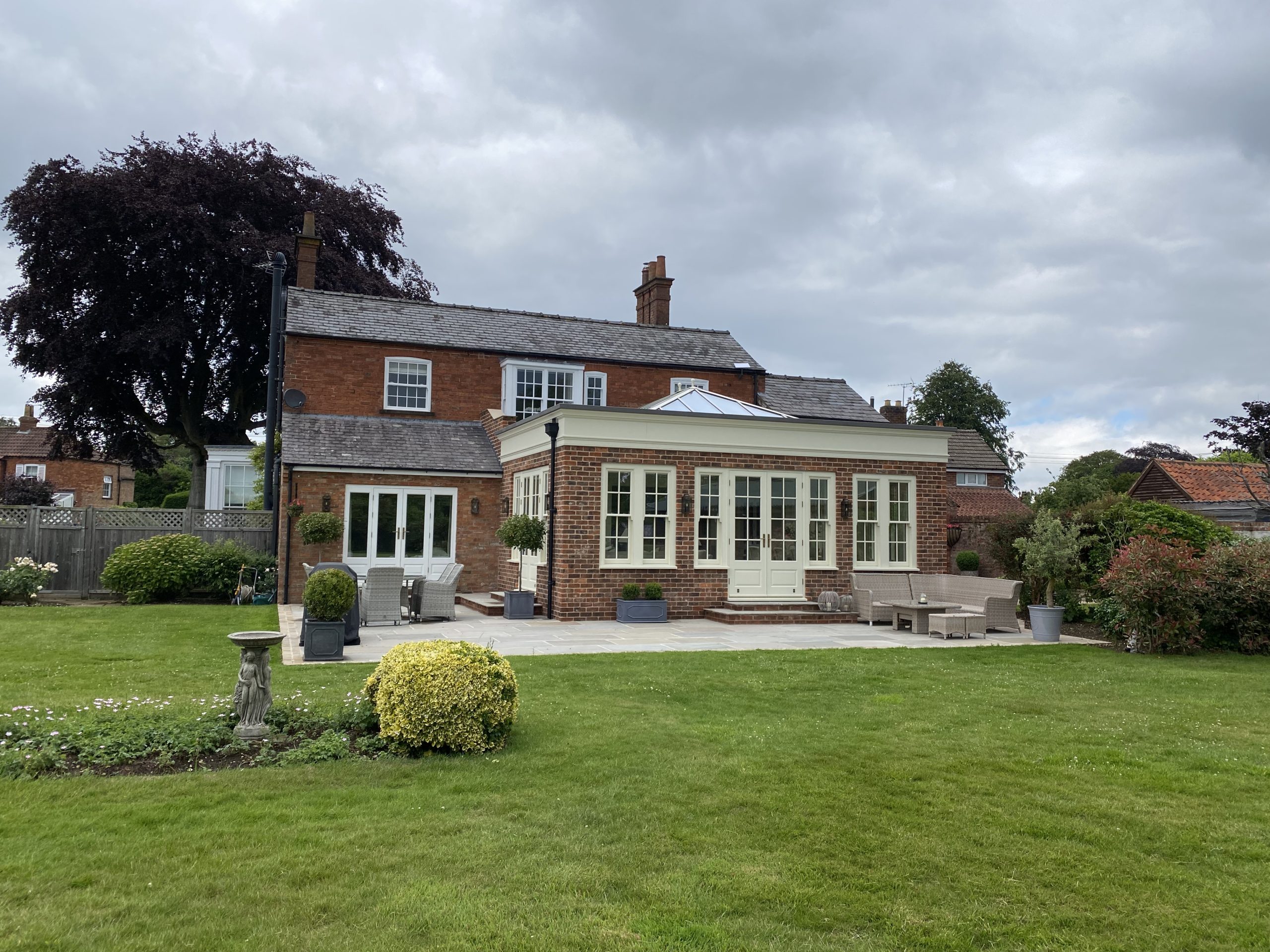 A large brick house with white-framed windows and an attached conservatory sits behind a well-kept lawn and garden on a cloudy day.